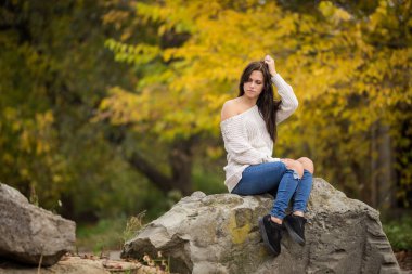 Güzel bir kız saçlarını düzeltiyor. Güz parkında gri bir taşın üzerinde oturuyor. Sarı yaprakların arka planında.