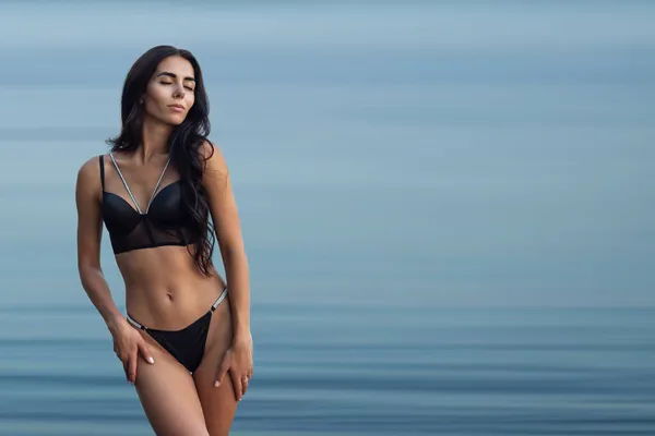Ung sexig brunett i svart bikini som poserar mot bakgrunden av vatten, copyspace — Stockfoto