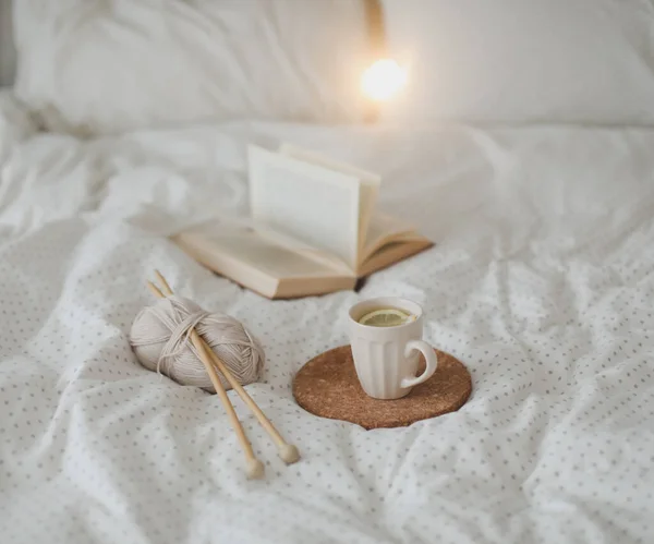 Cozy still life interior details with a book, tea cup and knitting needles in warm soft bed. Sweet home. — Stock Photo, Image
