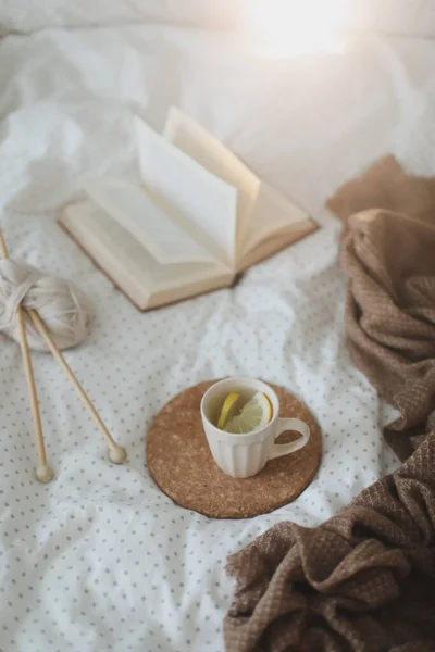 Cozy composition with a book, tea cup and knitting needles on bed sheets. Sweet home. — Stock Photo, Image