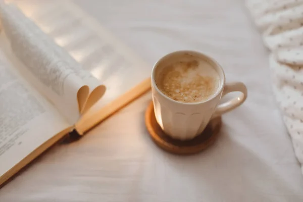 Composição de manhã acolhedora com uma xícara de café e um livro com páginas dobradas em forma de coração na cama — Fotografia de Stock