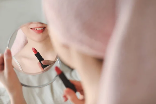 Красивая привлекательная улыбающаяся женщина красит губы красной помадой с помощью маленького круглого зеркала во время домашнего макияжа утром. — стоковое фото