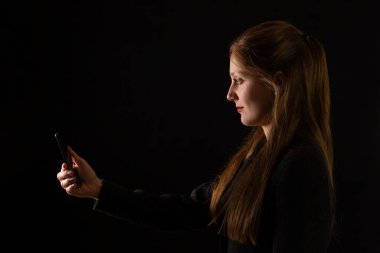 Young Caucasian woman holding smartphone in front of her face with black background. Side view portrait of female office worker interacting with modern technology at work.