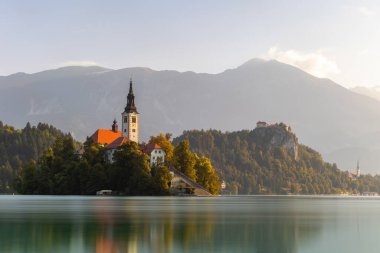 Nature scenery from Julien Alps with peak in background and church near water. Morning sun illuminating landscape with water, forest and sky in summer.