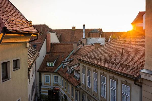 Sun setting over rooftops with red tiles in a city with old houses. Evening among buildings with shingles laid in regular pattern in Europe. High view of real estate properties in nice light.