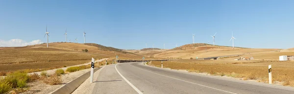 Beautiful panoramic photo of road through agrarian mountain landscape with windmills. On the road. Blue cloudy sky. Golden fields. Amazing scenery. Andalusia. Spain.