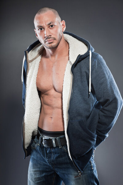 Muscled fitness man. Cool looking. Tough guy. Brown eyes. Bald. Wearing blue hoody shirt. Tanned skin. Studio shot isolated on grey background.