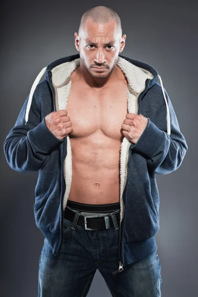 Homem musculado. Boa aparência. Um tipo duro. Olhos castanhos. Careca. Vestindo camisa azul com capuz. Pele bronzeada. Estúdio tiro isolado em fundo cinza . — Fotografia de Stock