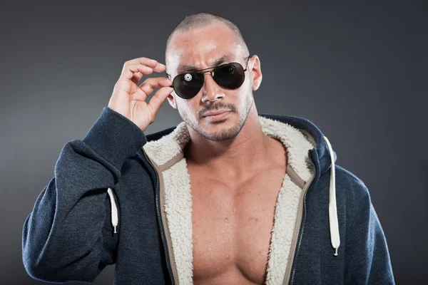 Muscled fitness man. Cool looking. Tough guy. Brown eyes. Bald. Wearing blue hoody shirt and sunglasses. Tanned skin. Studio shot isolated on grey background. — Stock Photo, Image