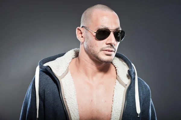 Muscled fitness man. Cool looking. Tough guy. Brown eyes. Bald. Wearing blue hoody shirt and sunglasses. Tanned skin. Studio shot isolated on grey background. — Stock Photo, Image