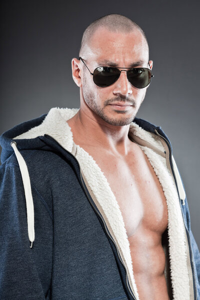Muscled fitness man. Cool looking. Tough guy. Brown eyes. Bald. Wearing blue hoody shirt and sunglasses. Tanned skin. Studio shot isolated on grey background.