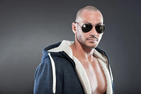 Muscled fitness man. Cool looking. Tough guy. Brown eyes. Bald. Wearing blue hoody shirt and sunglasses. Tanned skin. Studio shot isolated on grey background. — Stock Photo, Image