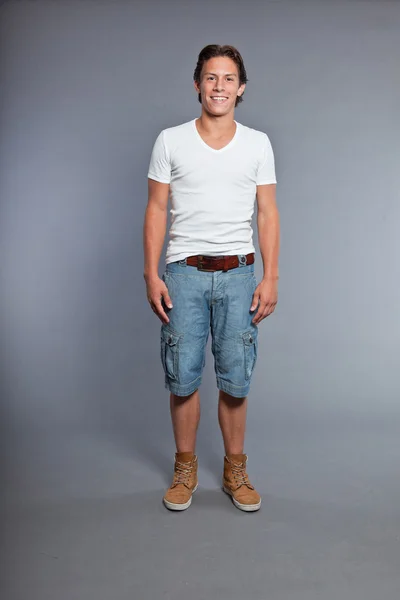 Ragazzo con i capelli castani e gli occhi. Indossa maglietta bianca e pantaloncini blu. Bell'aspetto. Abbigliamento casual. Espressioni. Studio ritratto isolato su sfondo grigio . — Foto Stock