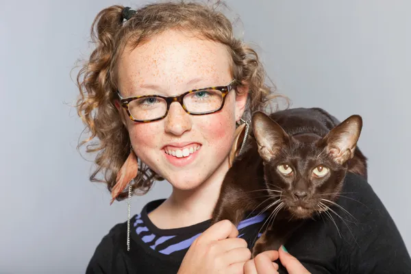 Gelukkig tienermeisje met glazen en blond krullend haar knuffelen donker bruin Oosterse shorthait kat. — Stockfoto