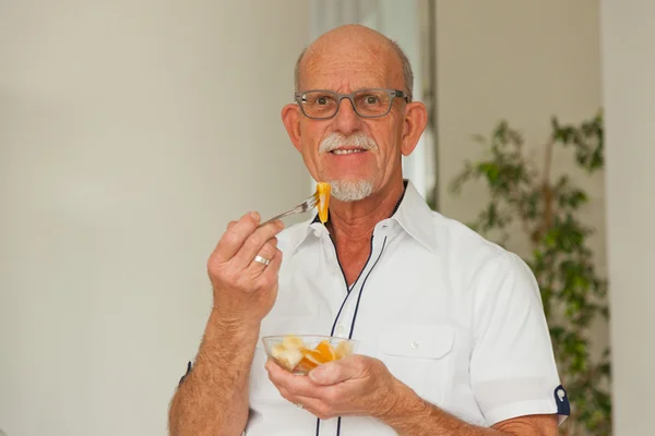 Senior man eten van verse fruitmand. vergadering in woonkamer. — Stockfoto