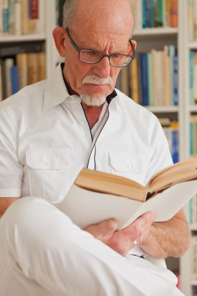 Äldre man med glasögon läser bok framför bokhylla. — Stockfoto