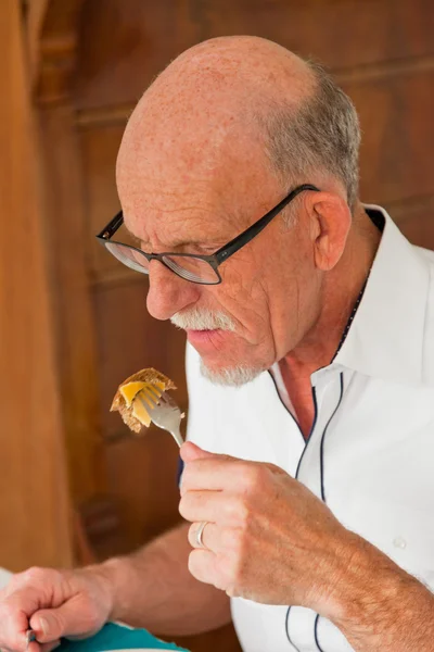 Senior man with glasses eating bread with cheese at table. — Stock Photo, Image