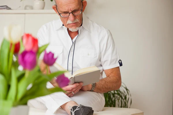 Senior man with glasses reading book in living room. — Stock Photo, Image