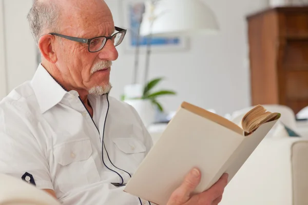 Senior man met bril lezen boek in woonkamer. — Stockfoto