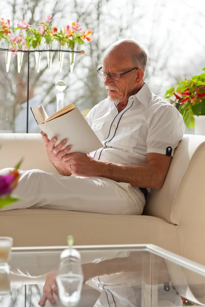 Senior man with glasses reading book in living room. — Stock Photo, Image