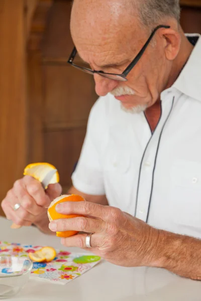 Hombre mayor con gafas pelando naranja fresca. Sentado en la mesa . —  Fotos de Stock
