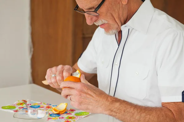 Homem sênior com óculos descascando laranja fresca. Sentado à mesa . — Fotografia de Stock