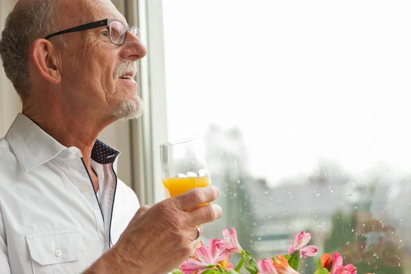 Senior man with glasses drinking orange juice at window with flo — Stock Photo, Image
