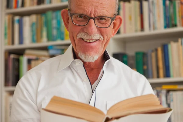 Äldre man med glasögon läser bok framför bokhylla. — Stockfoto