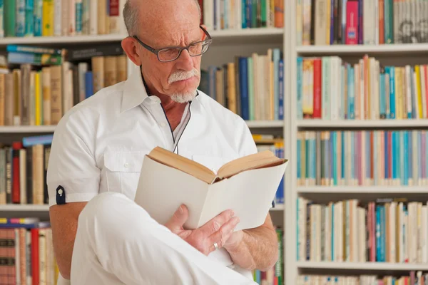 Senior mit Brille liest Buch vor Bücherregal. — Stockfoto