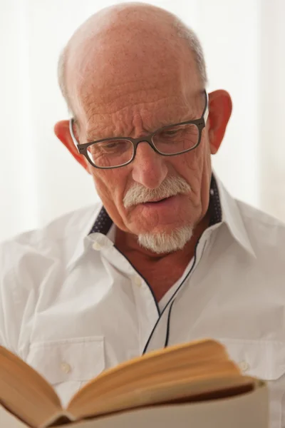 Senior man met bril lezen boek in woonkamer. — Stockfoto