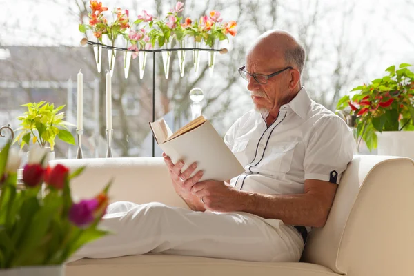 Senior man met bril lezen boek in woonkamer. — Stockfoto