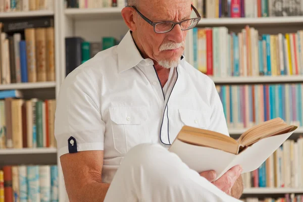 Senior mit Brille liest Buch vor Bücherregal. — Stockfoto