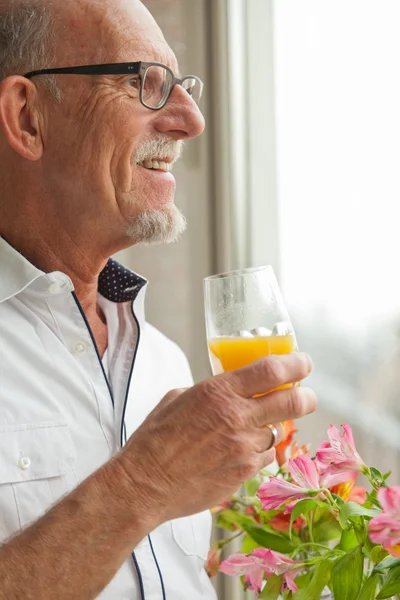 Homem sênior com óculos bebendo suco de laranja na janela com flo — Fotografia de Stock