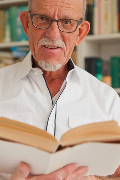 Hombre mayor con gafas leyendo libro delante de la estantería . — Foto de Stock