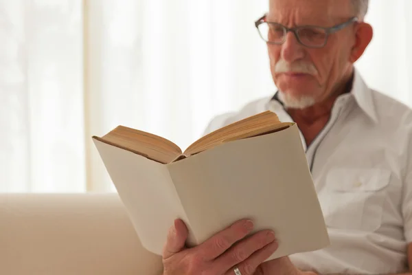 Senior man met bril lezen boek in woonkamer. — Stockfoto