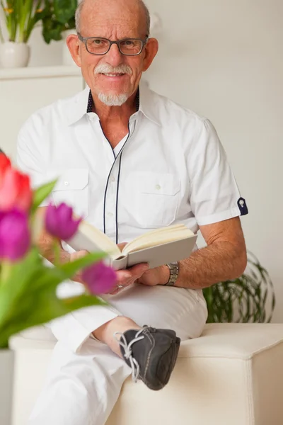 Senior man with glasses reading book in living room. — Stock Photo, Image