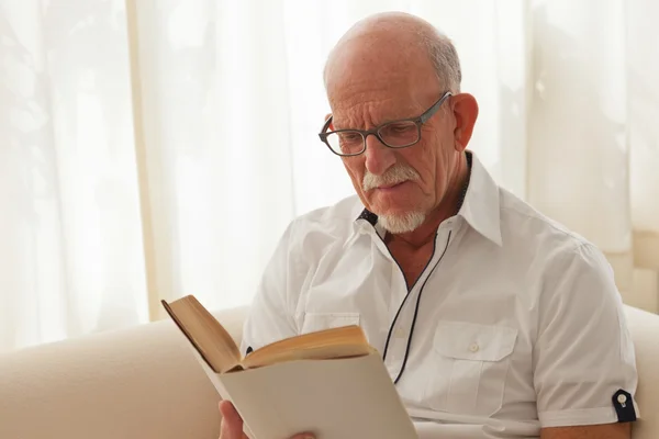 Äldre man med glasögon läsa bok i vardagsrummet. — Stockfoto