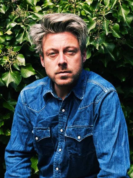 Man with blonde hair in front of green foliage. Wearing jeans sh — Stock Photo, Image