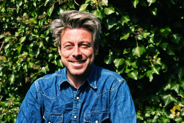 Man with blonde hair in front of green foliage. Wearing jeans sh — Stock Photo, Image