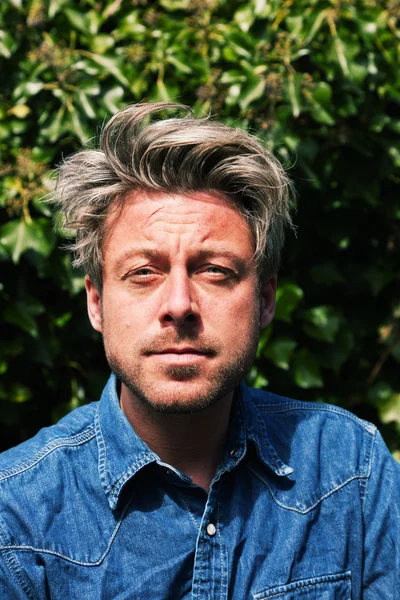 Man with blonde hair in front of green foliage. Wearing jeans sh — Stock Photo, Image