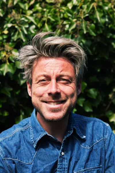 Man with blonde hair in front of green foliage. Wearing jeans sh — Stock Photo, Image