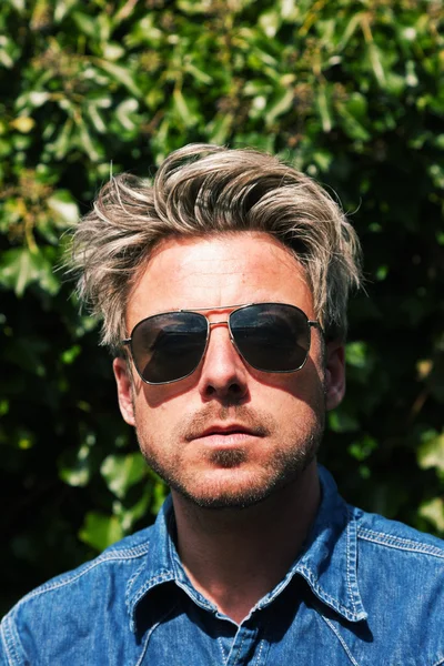 Homme aux cheveux blonds devant un feuillage vert. Porter des lunettes de soleil — Photo