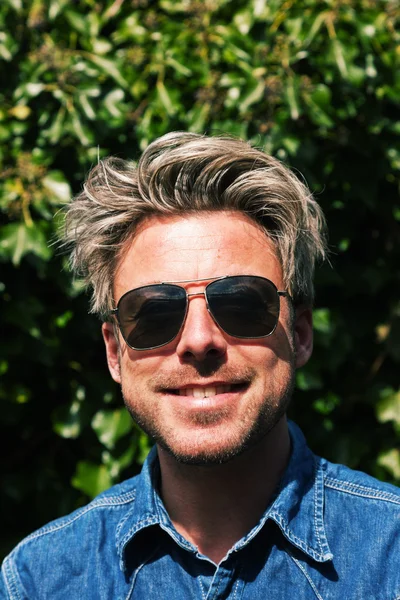 Mann mit blonden Haaren vor grünem Laub. mit Sonnenbrille — Stockfoto