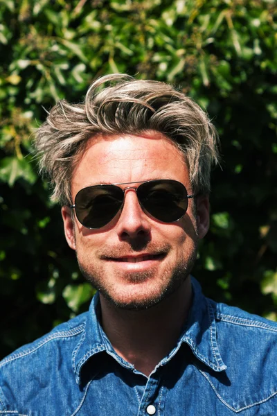 Mann mit blonden Haaren vor grünem Laub. mit Sonnenbrille — Stockfoto