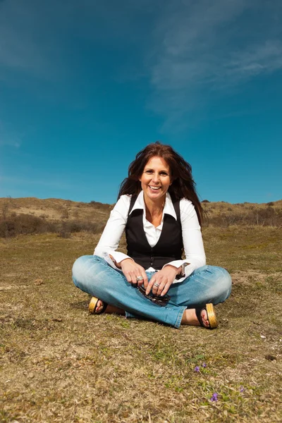 Smiling pretty brunette woman wearing jeans sitting in meadow wi — Stock Photo, Image