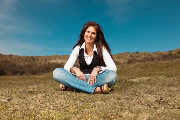 Hymyilevä kaunis brunette nainen yllään farkut istuu niitty wi — kuvapankkivalokuva