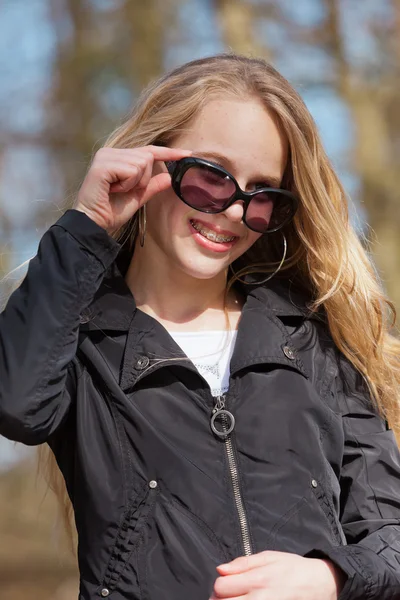 Leende ung blond flicka med solglasögon och hängslen i park. — Stockfoto