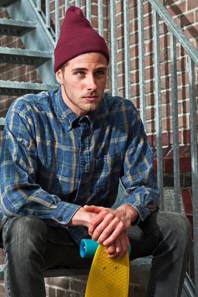 Urban skateboarder with woolen hat holding his skateboard sittin — Stock Photo, Image