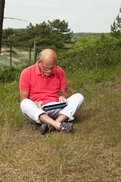 Senior retired man sitting with tablet outdoors in meadow. Scott — Stock Photo, Image