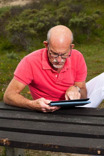 Homem idoso aposentado descansando e usando seu tablet à mesa no parque — Fotografia de Stock